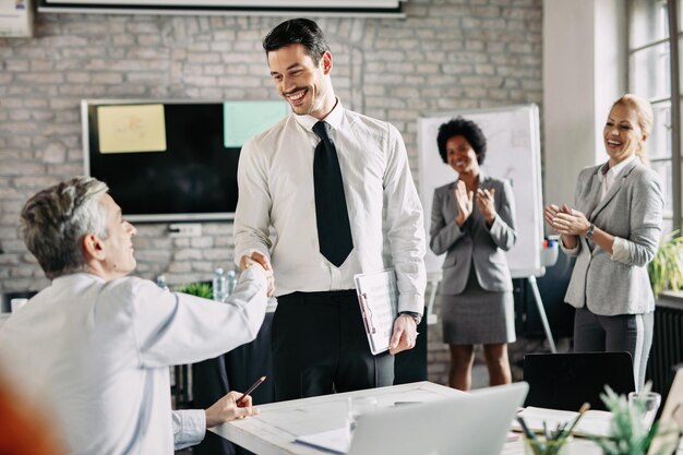 Two happy businessmen shaking hands after successful agreement in the office while their female colleagues are applauding them Focus is on young businessman