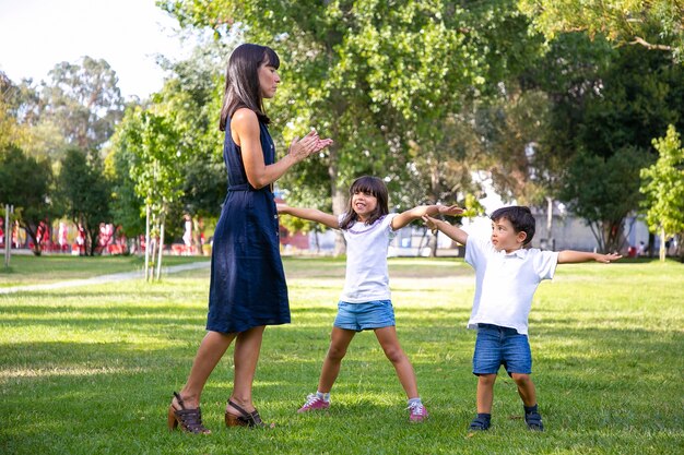 Two happy cute kids and their mom playing active games outdoors, doing exercises on grass in park. Family outdoor activity and leisure concept