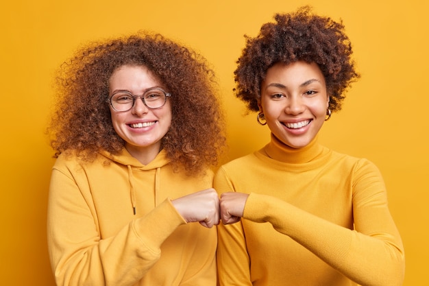 Free Photo two happy diverse women make fist bumps demonstrate agreement have friendly relationship smile gladfully stand next to each other isolated over yellow wall. teamwork body language concept