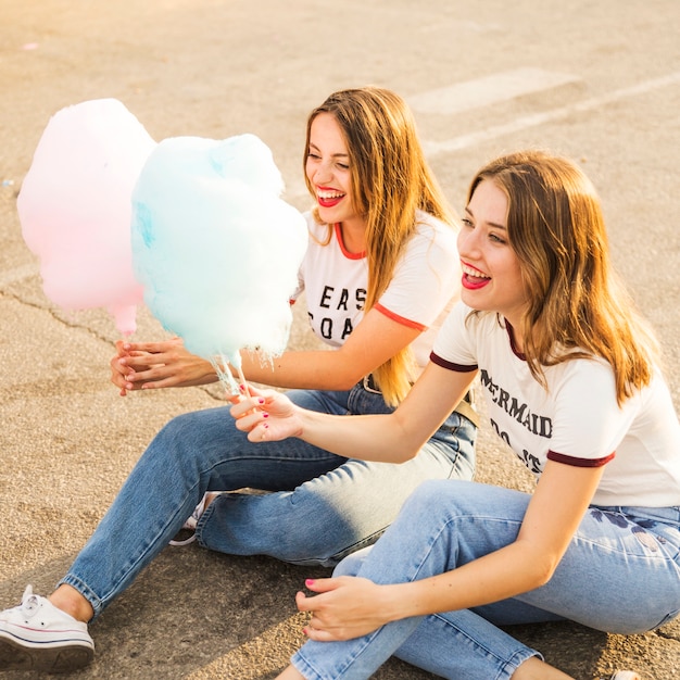 Free photo two happy female friends sitting on street holding candy floss