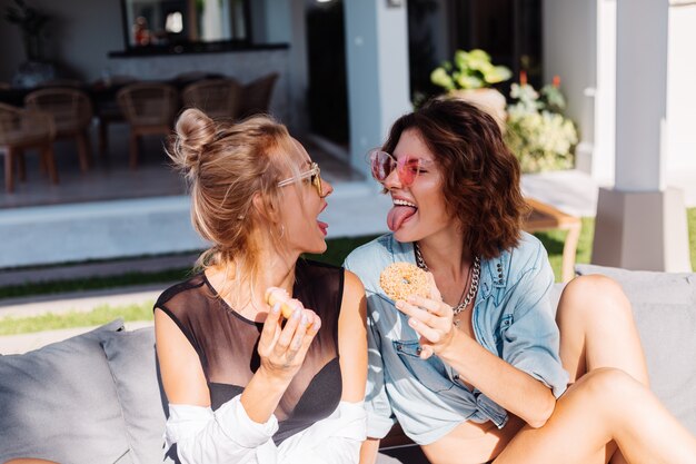 Two happy fit woman in pink and yellow sunglasses smiling having fun laughing with donuts, outdoor