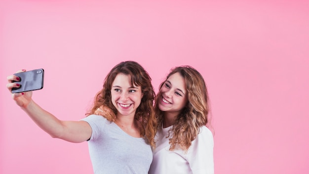 Two happy friends taking self portrait on cellphone standing against pink backdrop