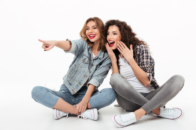 Two happy girls sitting on the floor together and looking away over white wall