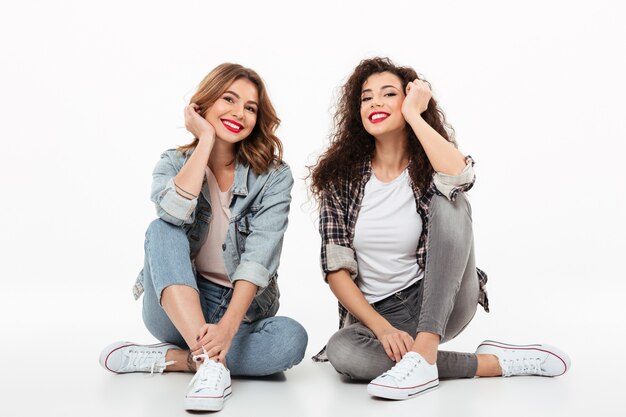 Two happy girls sitting on the floor together  over white wall