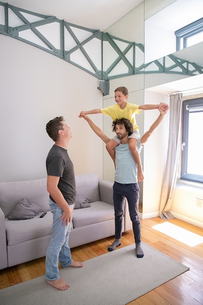 Two happy homosexual dads and son having fun at home, boy riding on mans neck and laughing. Full length. Family and parenthood concept