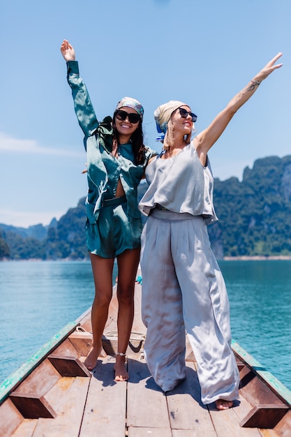 Two happy woman blogger tourist friends in silk suit and scarf and sunglasses on vacation travel around thailand on asian boat, Khao Sok national park.