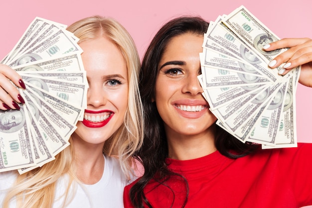 Two happy women covering their half faces and looking at the camera over pink 