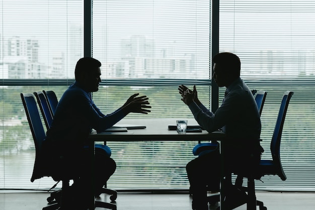 two human outlines against the shuttered office window sitting opposite each other and negotiating