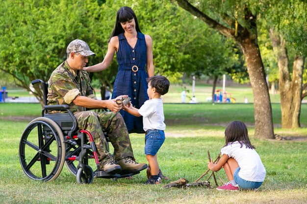 Two kids arranging wood for campfire outdoors near mom and disabled military dad in wheelchair. Boy showing log to father. Disabled veteran or family outdoors concept