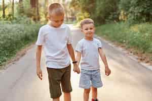 Free photo two little boys wearing white t shirts and shorts going together and holding hands in summer park, brothers walking outdoor, expressing positive emotions.