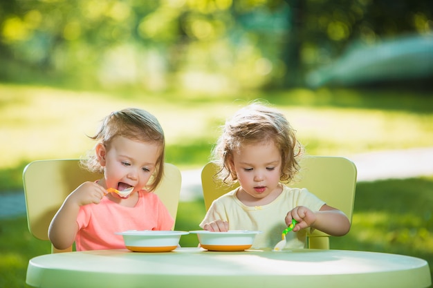 Two little girls sitting at a table and eating together against green lawn