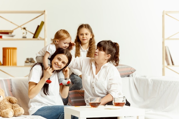 Two little girls their attractive young mother and their charming grandmother sitting on sofa and spending time together at home. Generation of women. International Women's Day. Happy Mother's Day.