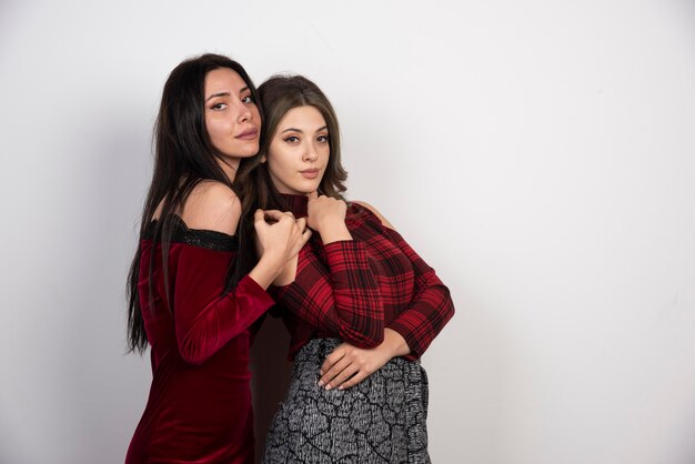 Two lovely attractive girls standing and looking at camera on white wall.