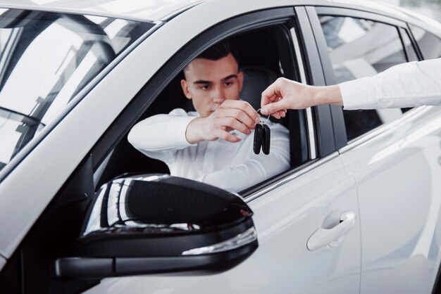 Two men stand in the showroom against cars. Close-up of a sales manager in a suit that sells a car to a customer. The seller gives the key to the customer.