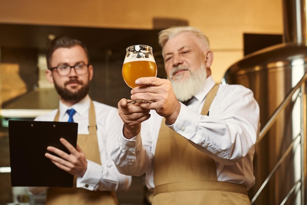 Two men wearing keeping glass of beer and examining quality