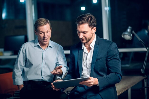 Two office workers with tablet in office late at night. Young businessmen talking while viewing presentation on digital touchscreen tablet.