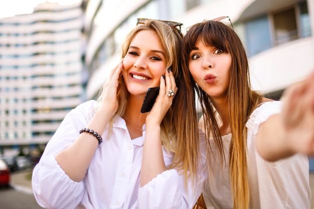 Two pretty blonde wand woman positive skills sisters woman making selfie in the street, summer sunny colors, white shirts exited emotions.