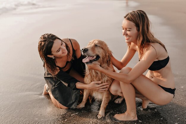 Free Photo two pretty girls on the beach near the ocean playing with a dog on sunset