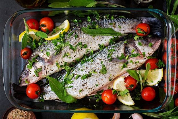 Two raw seabass in a baking dish with spices on an black table. Top view