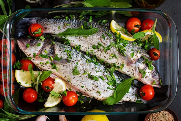 Two raw seabass in a baking dish with spices on an black table. Top view