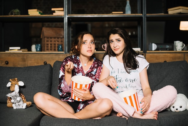 Free photo two scared women sitting on sofa with popcorn watching television