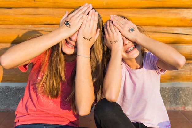 Free Photo two sisters sitting outside covering their eyes with eyes tattoo on palm