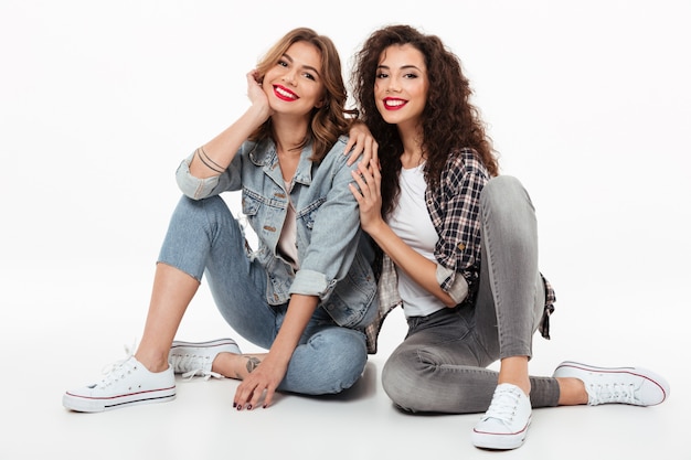 Free Photo two smiling girls sitting on the floor together  over white wall