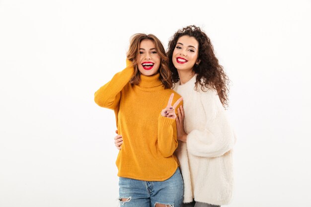 Two smiling girls in sweaters posing together  while one woman showing peace gesture over white wall