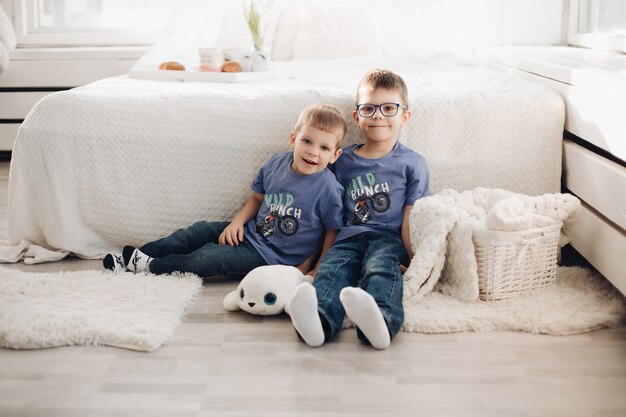 Two smiling male kid posing together at comfortable white bedroom interior. Happy brothers hugging having fun at cosiness home sitting on floor near bed