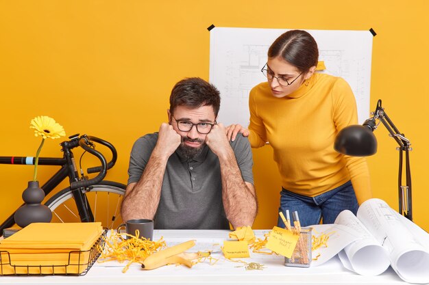 Two students prepare for upcoming examination sit at office desk with papers blueprints and sketches. Frustrated unhappy bearded man feels tired after preparing architectural project. Teamworking