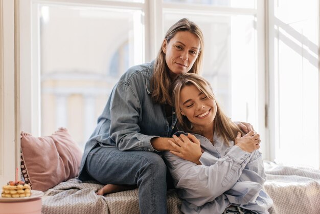 Two stylish caucasian blonde women in casual clothes smile and hug while sitting at window during day. Love expression concept