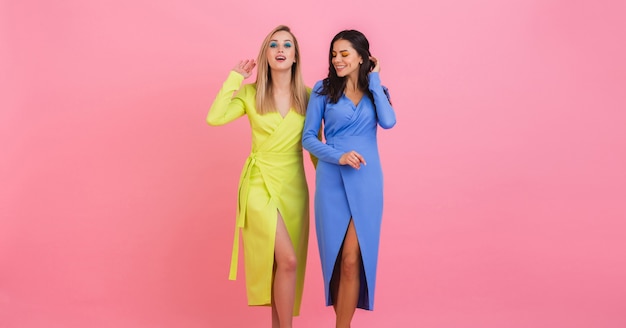 Two stylish smiling attractive women friends posing on pink wall in stylish colorful dresses of blue and yellow color, spring fashion trend