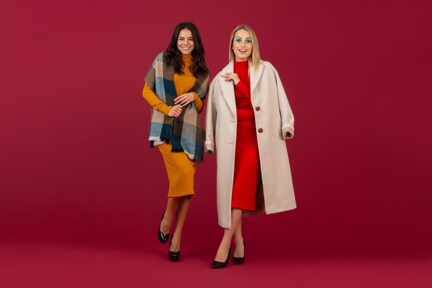 Two stylish women in autumn winter fashion dress and coat posing isolated  on red wall