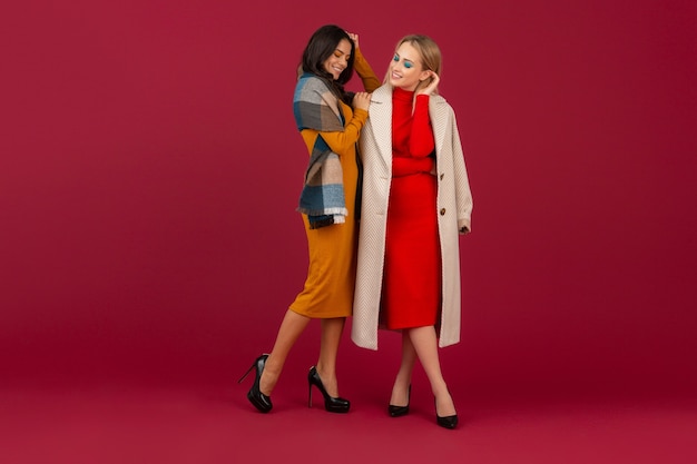 Two stylish women in autumn winter fashion dress and coat posing isolated  on red wall 