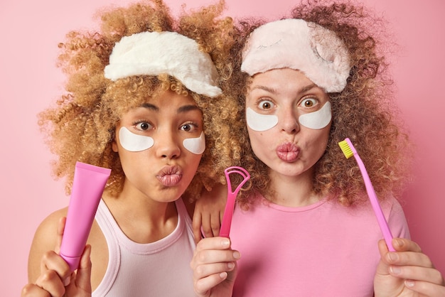 Free Photo two surprised female sisters with curly hair wears sleepmasks apply beauty patches under eyes going to clean teeth and tongue with brush stand closely to each other isolated over pink background