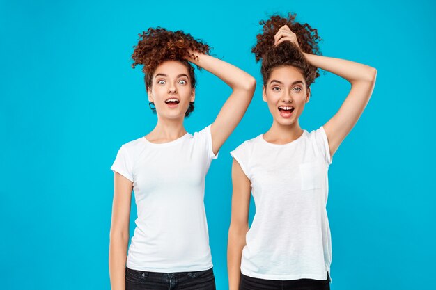 Two surprised womans twins holding hair, joking over blue.