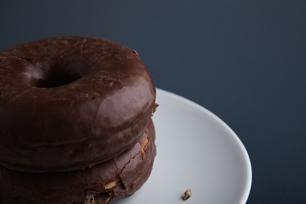 Two tasty freshly baked donuts glazed with chocolate on white ceramic small plate isolated in corner of rustic old blue wooden table. Close up focus with detail