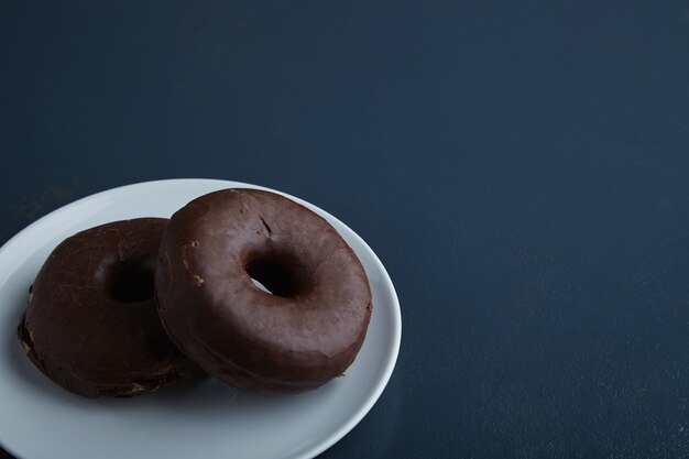 Two tasty freshly baked donuts glazed with chocolate on white ceramic small plate isolated in corner of rustic old blue wooden table