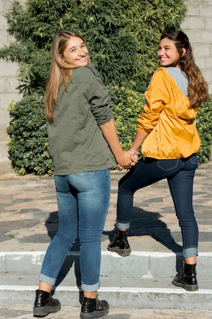 Two teenage girls holding each other's hand looking over shoulder at outdoors