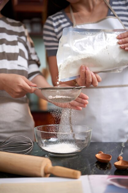 Two unrecognizable women pouring flour into sieve in kitchen at home