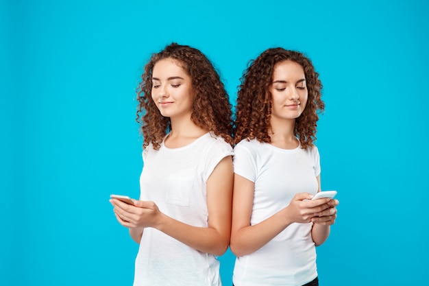 Free photo two womans twins smiling, looking at phones over blue.