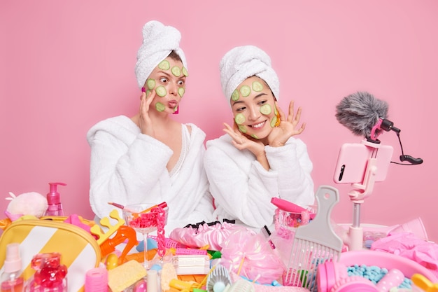 Free photo two women bloggers show how to make natural face mask apply cucumber slices on face and record vlog video on smartphone wear white soft bathrobes and towels over head isolated over pink wall