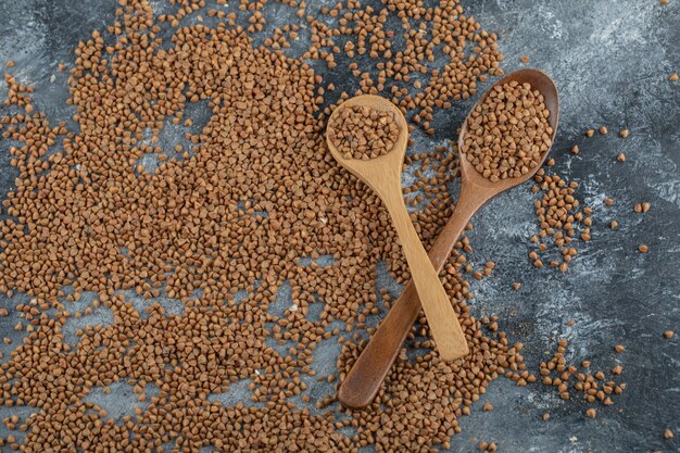 Two wooden spoons full of raw buckwheat on marble surface