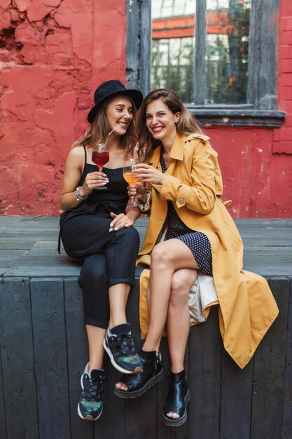 Two young attractive cheerful women with cocktails in hands while joyfully spending time together in old cozy courtyard of cafe