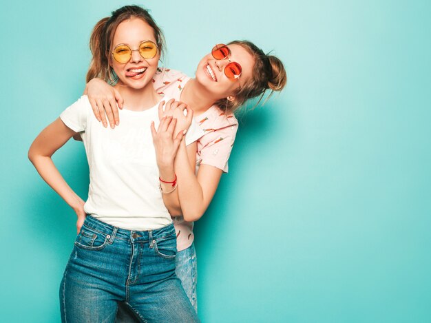 Two young beautiful blond smiling hipster girls in trendy summer hipster jeans clothes. Sexy carefree women posing near blue wall. Trendy and positive models having fun in sunglasses