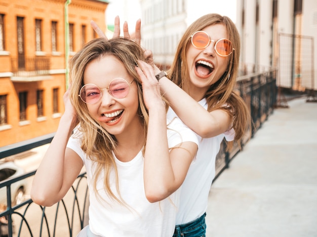 Free Photo two young beautiful blond smiling hipster girls in trendy summer white clothes.   .positive models having fun in sunglasses.using fingers as bunny ears