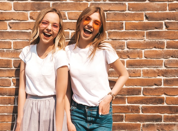 Two young beautiful blond smiling hipster girls in trendy summer white t-shirt clothes.  . Positive models having fun in sunglasses