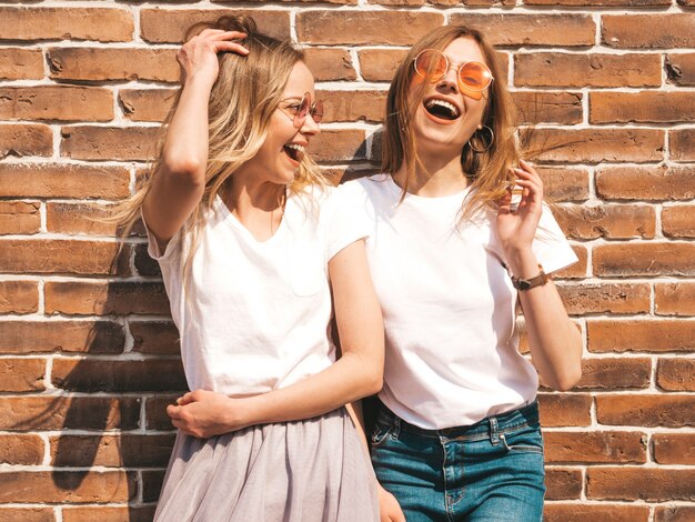 Two young beautiful blond smiling hipster girls in trendy summer white t-shirt clothes.  . Positive models having fun in sunglasses