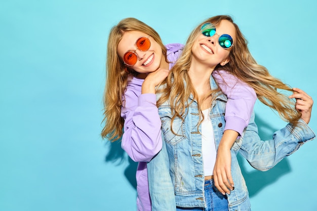 Two young beautiful blond smiling hipster women in trendy summer clothes. Sexy carefree women posing near blue wall in sunglasses. Positive models going crazy and hugging