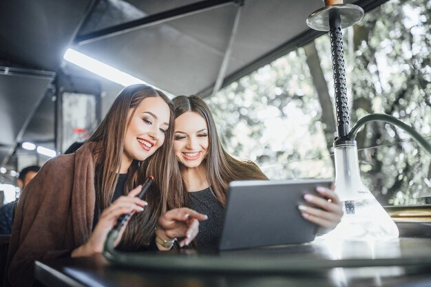 Two young beautiful girlfriends sitting on a summer terrace cafe, smoking hookah and looking at the tablet